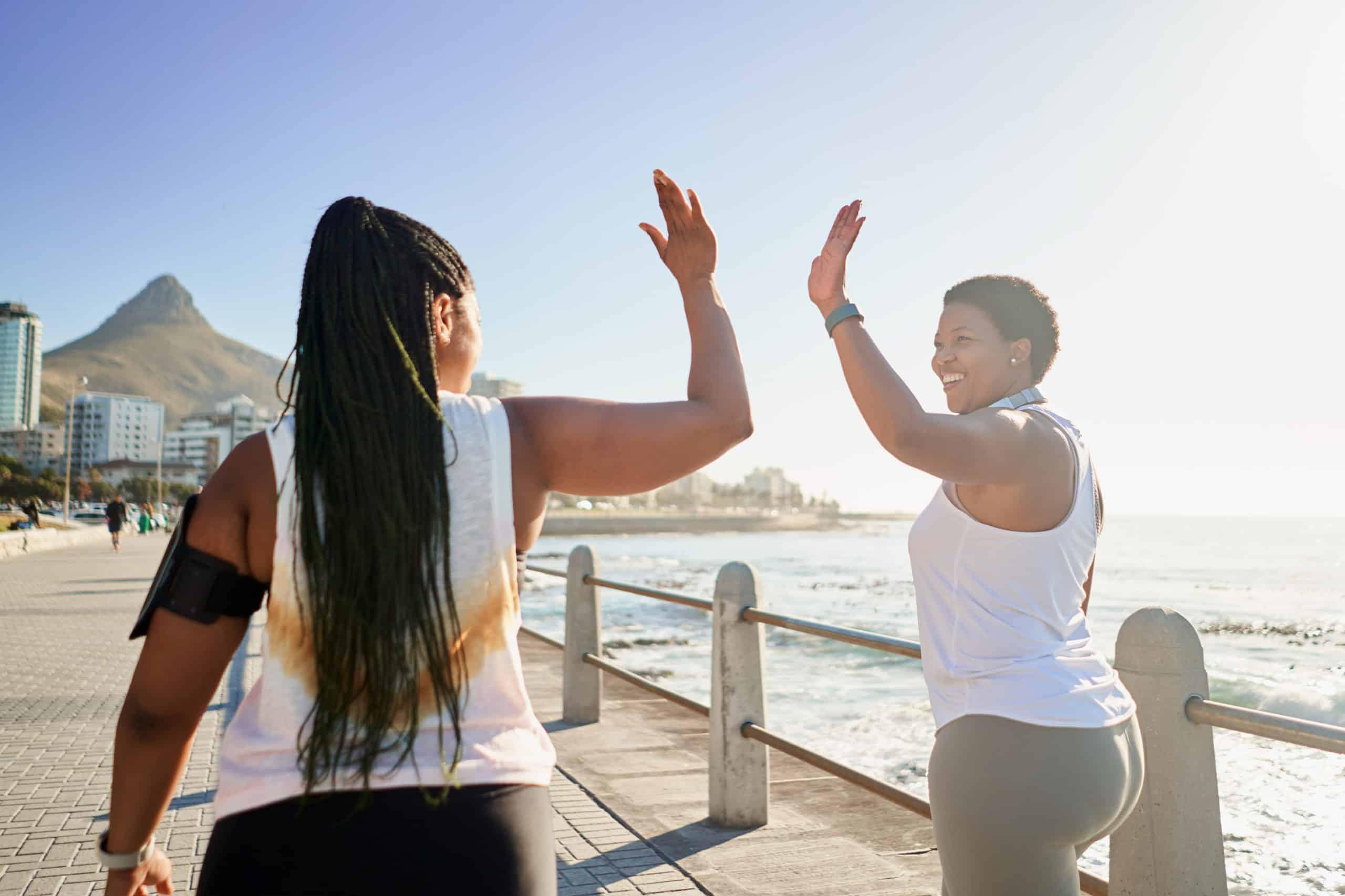 two Black fitness friends on a seaside boardwalk encourage and support each other during a summer outdoor workout. They are happy and confident and are wearing activity clothes and wearable tech.